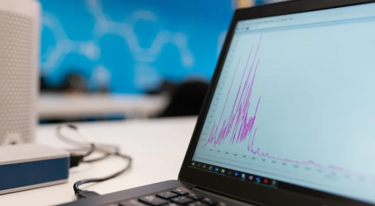 A laptop sits on a desk, open to display a graph with a pink line of data that jags up and down the screen. The graph is blurred to obfuscate the data.